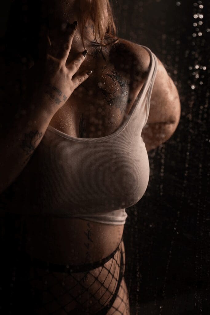 Woman in a white crop top and fishnet stockings posing behind a rain-covered glass panel during a moody shower scene boudoir session in Sioux Falls, South Dakota