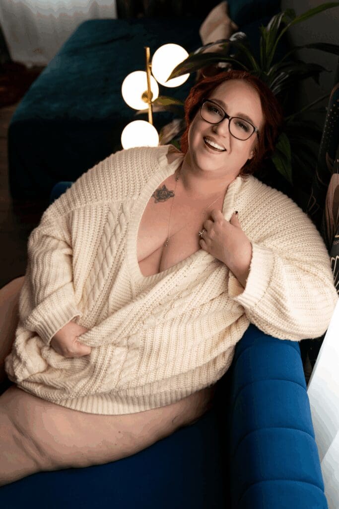 Smiling woman in an oversized cream sweater posing on a blue couch during a boudoir session in Sioux Falls, South Dakota